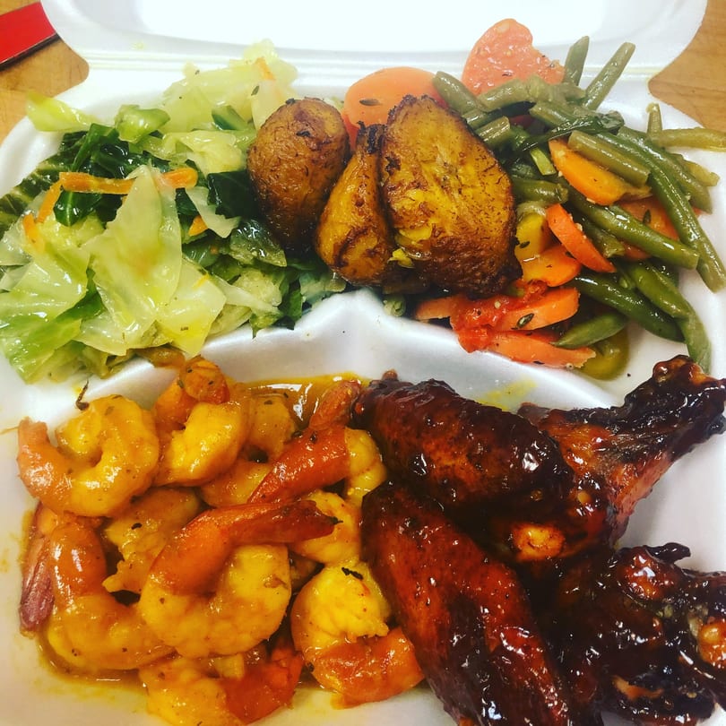 A Jamaican meal with jerk chicken, curried shrimp, plantains, cabbage, and green beans in a takeout container.