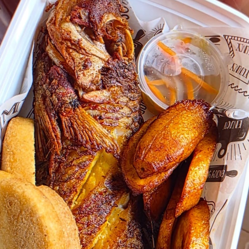 A Jamaican dish of fried fish, fried plantains, and a side of pickled vegetables in a container.